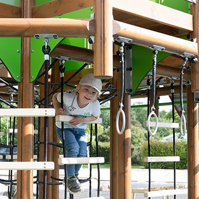 A little kid is climbing up a playground unit, looking out halfway the steps and looking into the camera smiling.